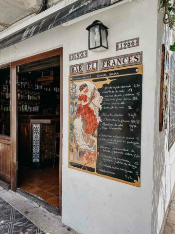 The entrance of Bar el Francés, featuring a charming vintage sign and a blackboard menu displaying various dishes. The exterior is decorated with plants and flowers, adding to the quaint and welcoming ambiance of the restaurant.