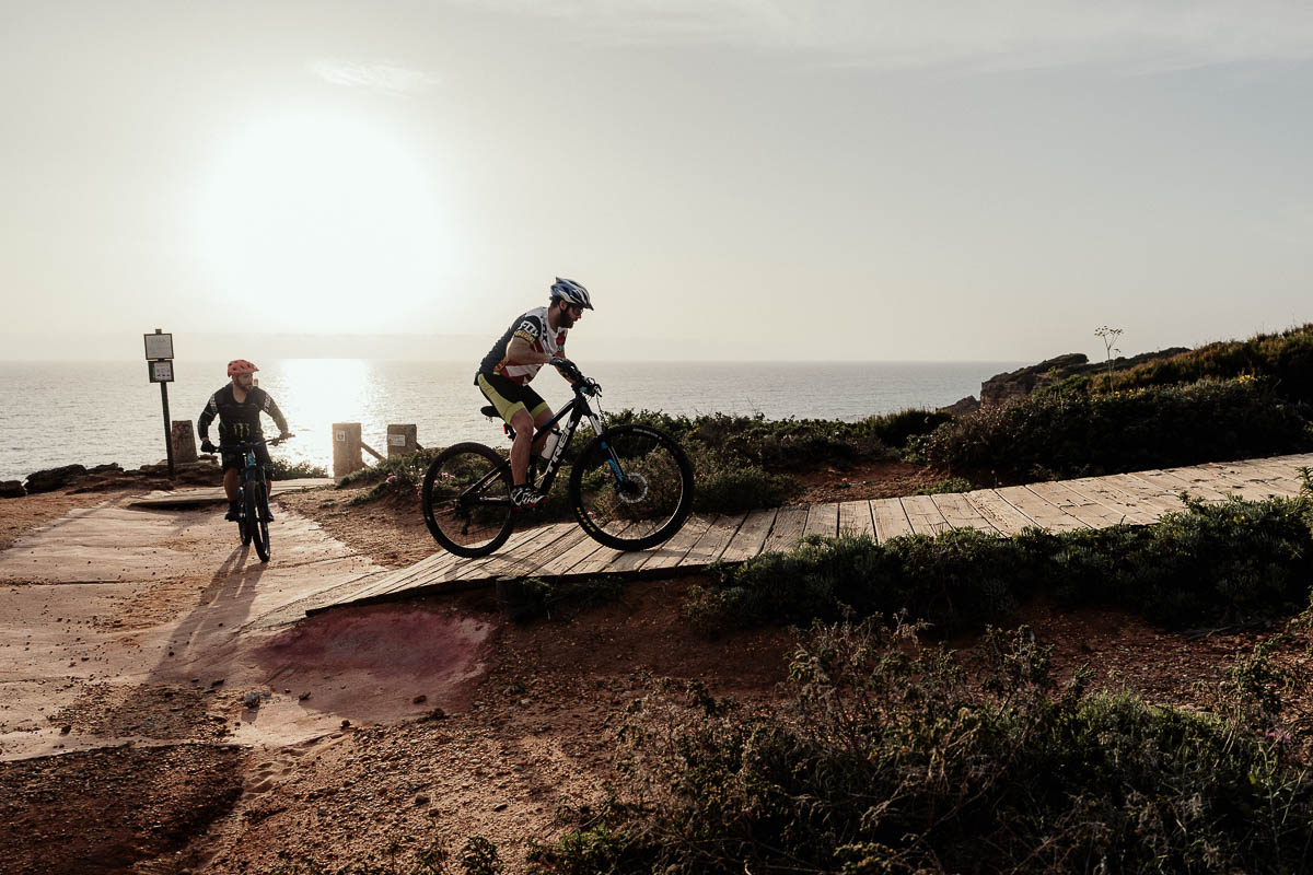 Two cyclists ride along a scenic pathway near the edge of a cliff, with the sun setting in the background. The golden light of the setting sun casts a warm glow over the landscape, highlighting the rugged terrain and the expansive ocean below. 