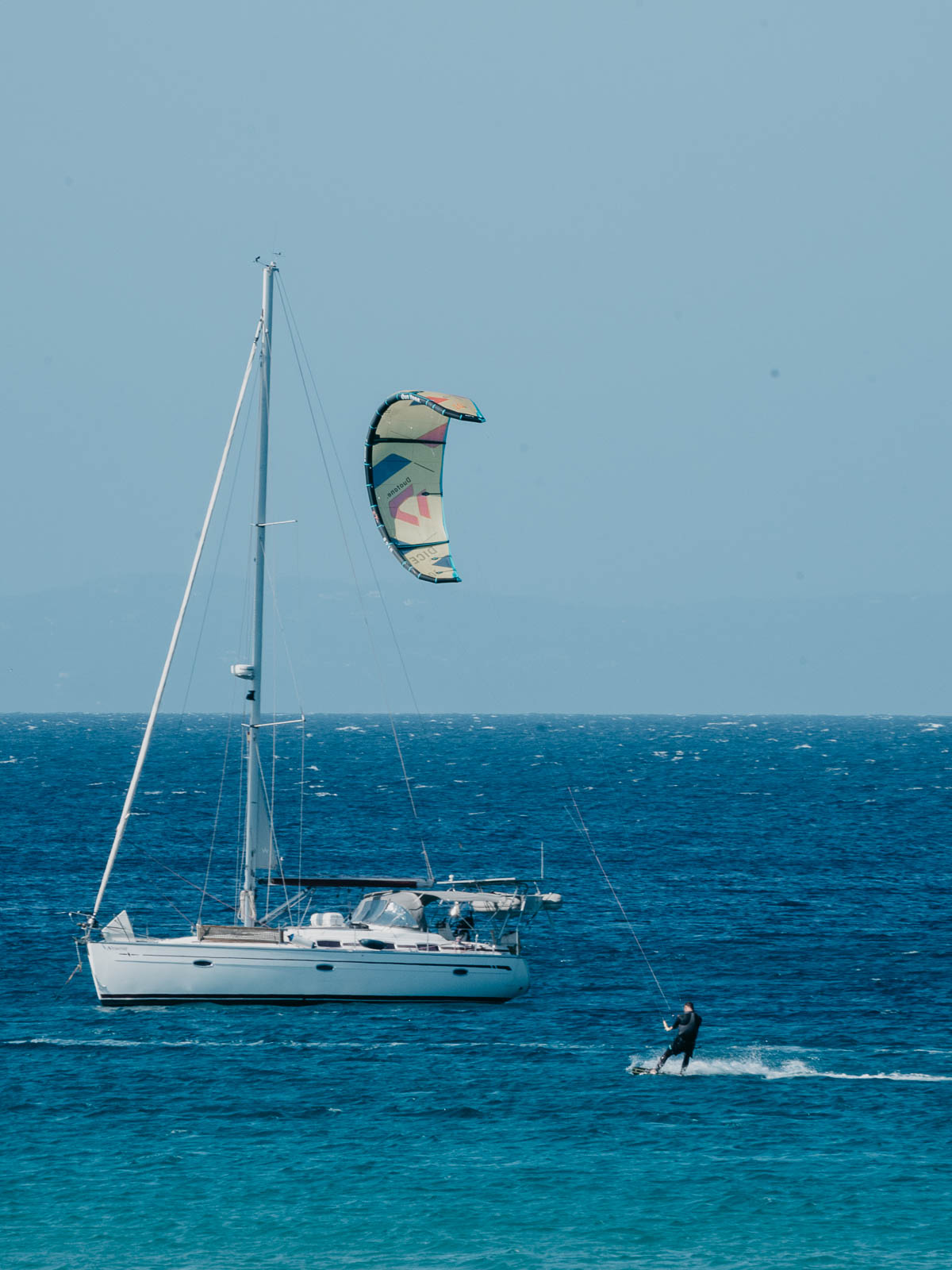 A sailboat is anchored on the calm sea with a kite surfer gliding nearby, creating a serene and adventurous scene against the clear blue water.