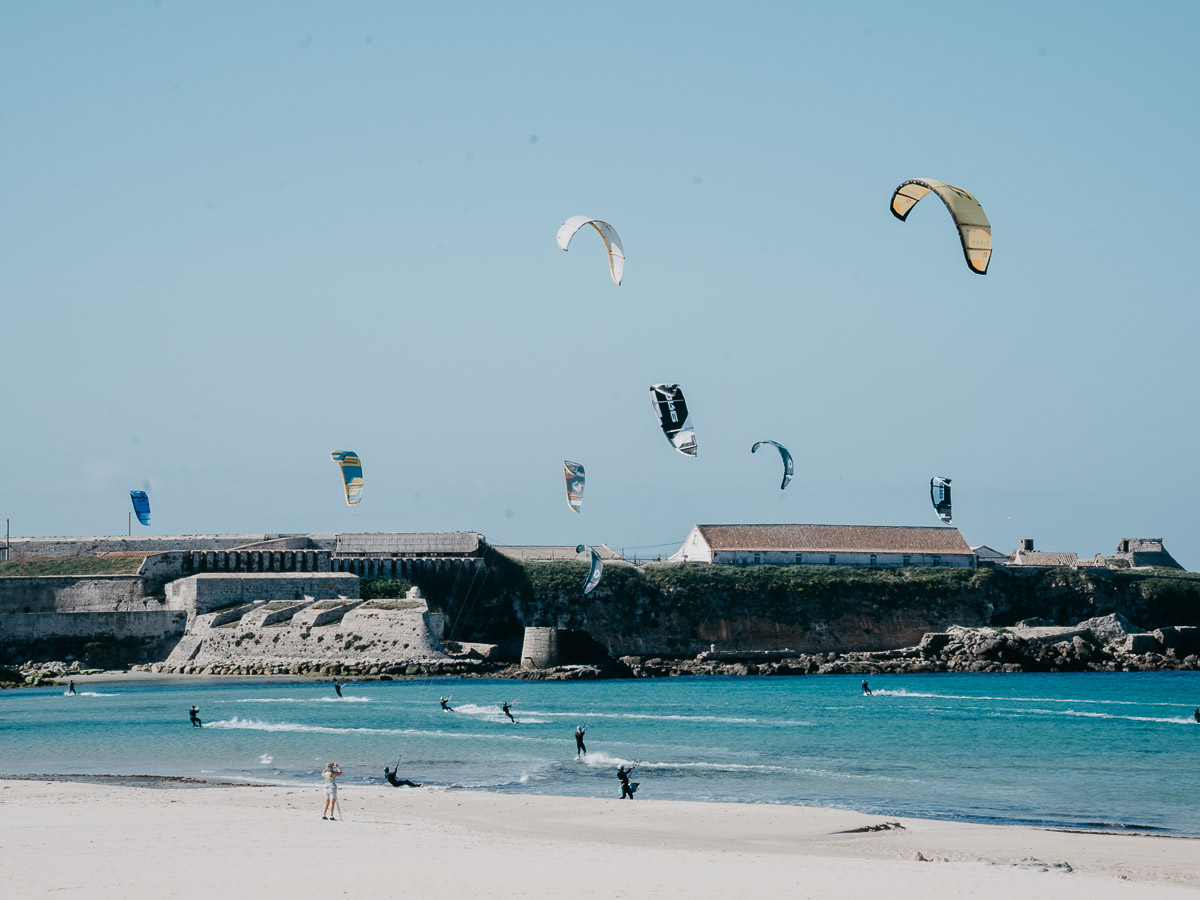 A coastal scene shows several kite surfers gliding across the water, their colorful kites soaring in the sky. In the background, a historic fortification stands on the rocky shore, contrasting with the bright blue sea and clear sky. 