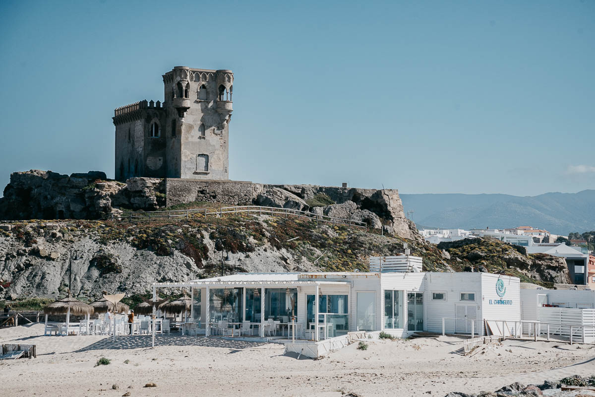 A beach scene featuring a historic stone tower standing on a rocky hilltop, overlooking a modern beachside restaurant named "El Chiringuito." The restaurant has a white, minimalist design with outdoor seating areas, contrasting with the ancient architecture of the tower. 