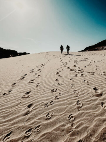 Two people walk across vast sand dunes under a bright sky, their footprints creating a trail behind them. The sun casts a dramatic light and shadow effect on the dunes, highlighting the texture of the sand and the sense of adventure in this natural setting.