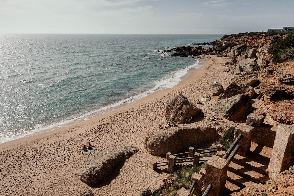 An aerial view of Calas de Roche, a secluded beach surrounded by dramatic rocky cliffs and crystal-clear turquoise waters. The sandy shore is dotted with large boulders and a few sunbathers, enjoying the tranquil and peaceful environment. The stairs leading down to the beach add a touch of rustic charm to this picturesque coastal spot.