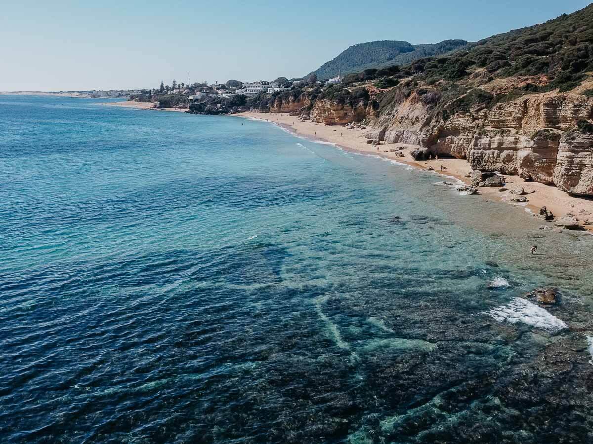 An aerial view of Caños de Meca, highlighting its stunning natural setting with dramatic cliffs, clear and blue waters.