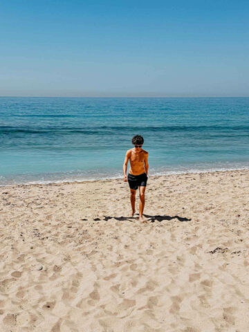 A person in swim trunks walking on the sandy shore of Caños de Meca towards the ocean, enjoying the serene and calm waters on a sunny day.