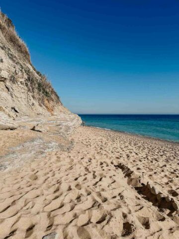 A view of a pristine sandy beach in Caños de Meca with dramatic cliffs on the left and clear blue waters stretching into the horizon under a bright blue sky.