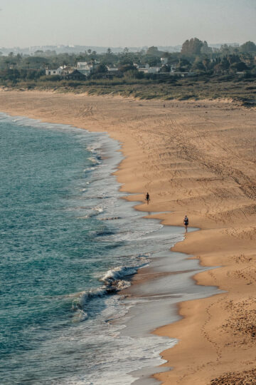 A long stretch of beach at Playa Faro de Trafalgar, with gentle waves lapping at the shore and a few people walking along the water's edge.