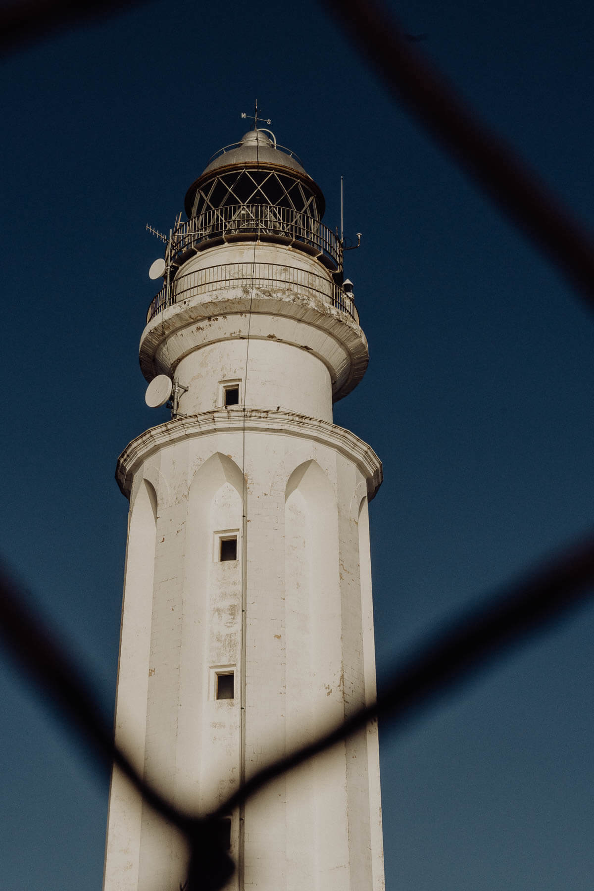 A close-up view of a historic white lighthouse against a deep blue sky, framed by a dark, blurred foreground element. 