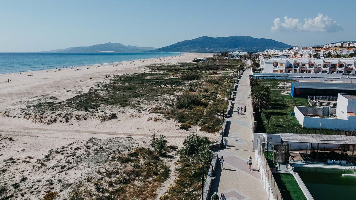 An aerial view of El Palmar de Vejer showcases a long, sandy beach stretching along the coastline, with a mix of natural vegetation and open spaces. On the right, a paved promenade runs parallel to the beach, lined with modern buildings and residential areas. In the distance, mountains and a clear blue sky provide a scenic backdrop.