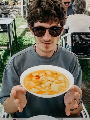 a man holds a bowl of hearty vegetable stew with white beans while seated outdoors.