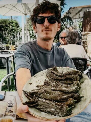 A man holds a plate of roasted green peppers, seated outdoors at the restaurant El Tropezón.