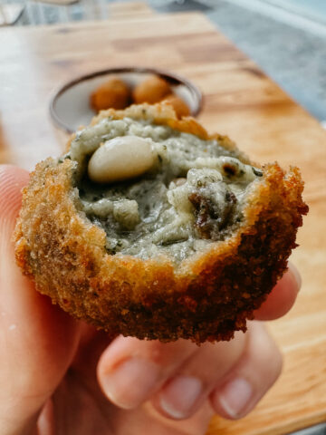 A close-up of a hand holding a croquette filled with mushrooms and pine nuts, showcasing its crispy exterior and savory filling. 