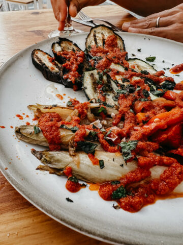 A plate of grilled vegetables topped with thick tomato sauce at El Lola. The dish includes eggplant, zucchini, and other veggies, garnished with herbs, showcasing a vibrant and appetizing vegan option.