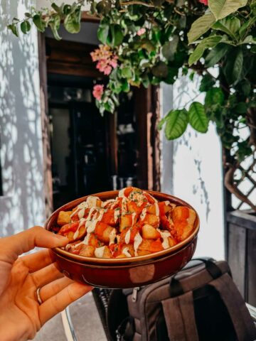 A hand holding a bowl of loaded patatas bravas topped with sauces and herbs. The background shows the entrance of Bar el Francés, adorned with green plants and flowers, providing a cozy and inviting atmosphere.