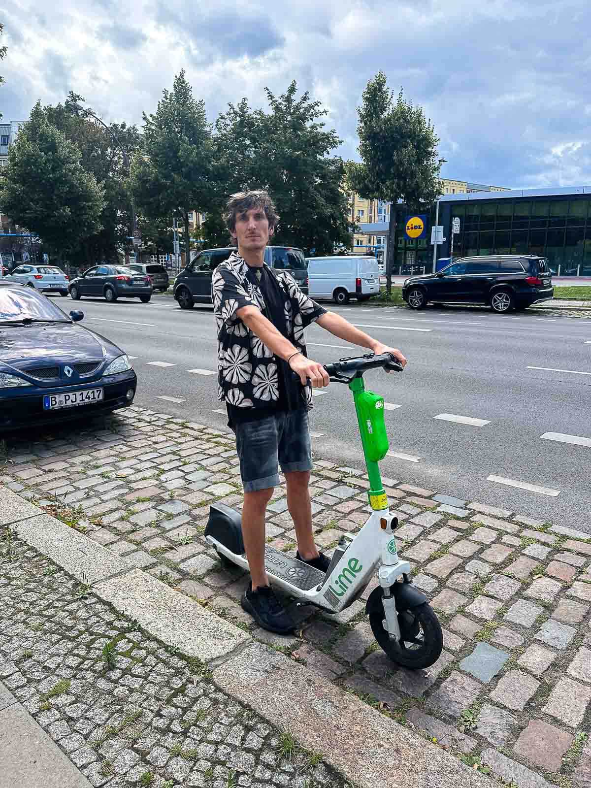 **Person Riding an Electric Scooter in Berlin**: A person standing on a green and white electric scooter on a cobblestone sidewalk in Berlin, with trees and cars visible in the background.