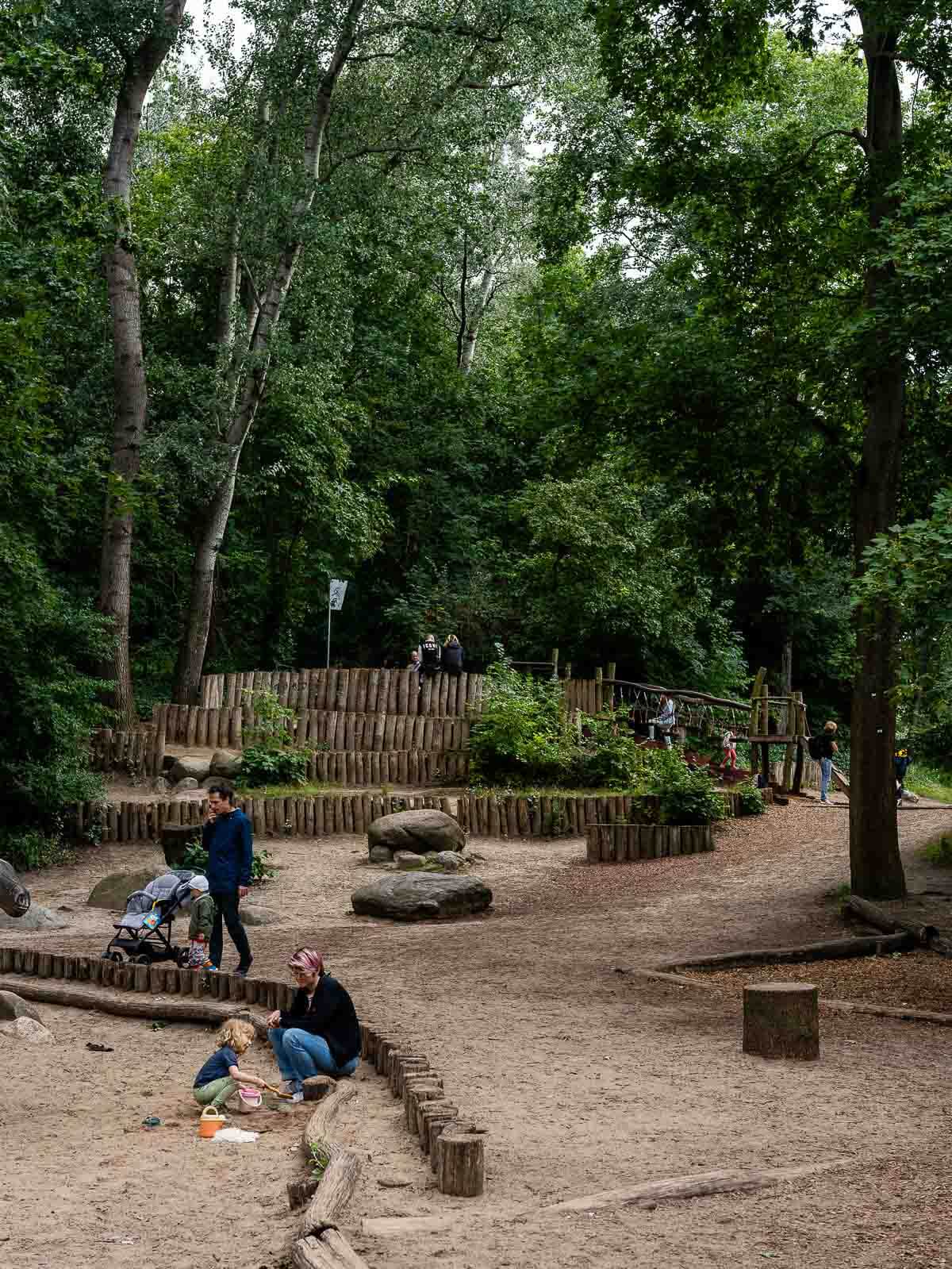 Children's Area in Park: Kids playing near a fenced area with various play equipment in a wooded park.