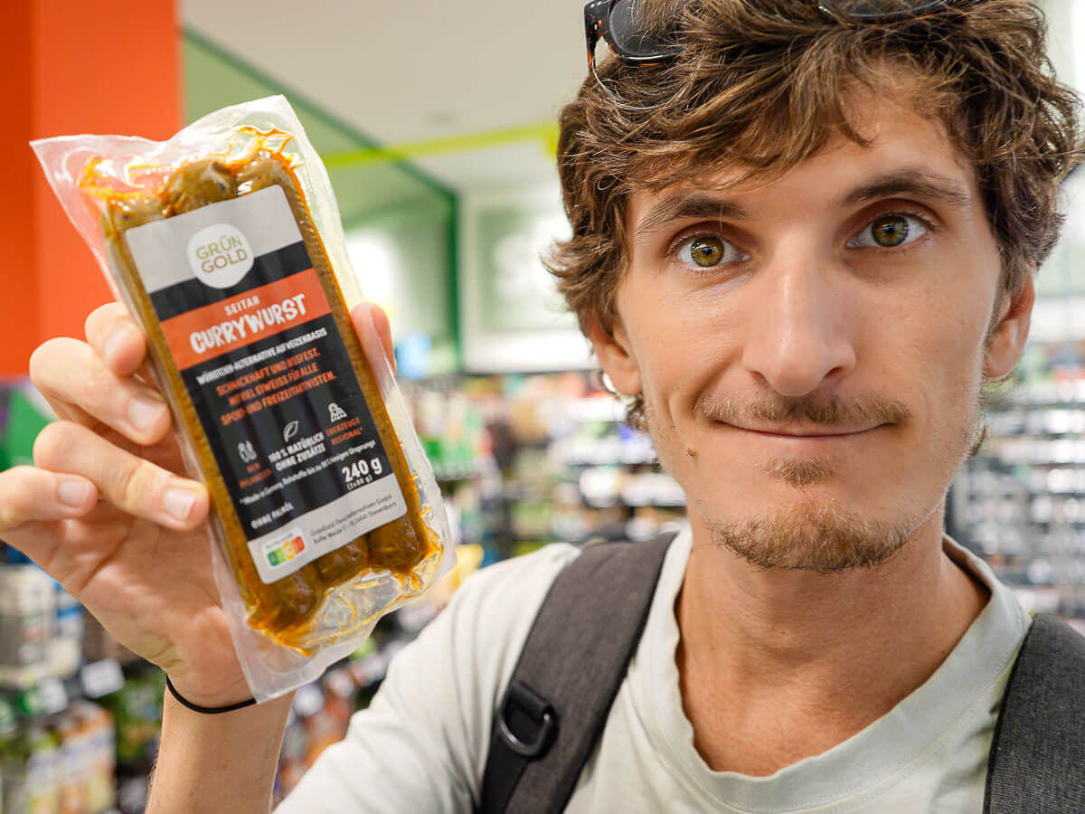 A close-up of a man holding a package of vegan sausages (currywurst) , showing it to the camera in a grocery store.