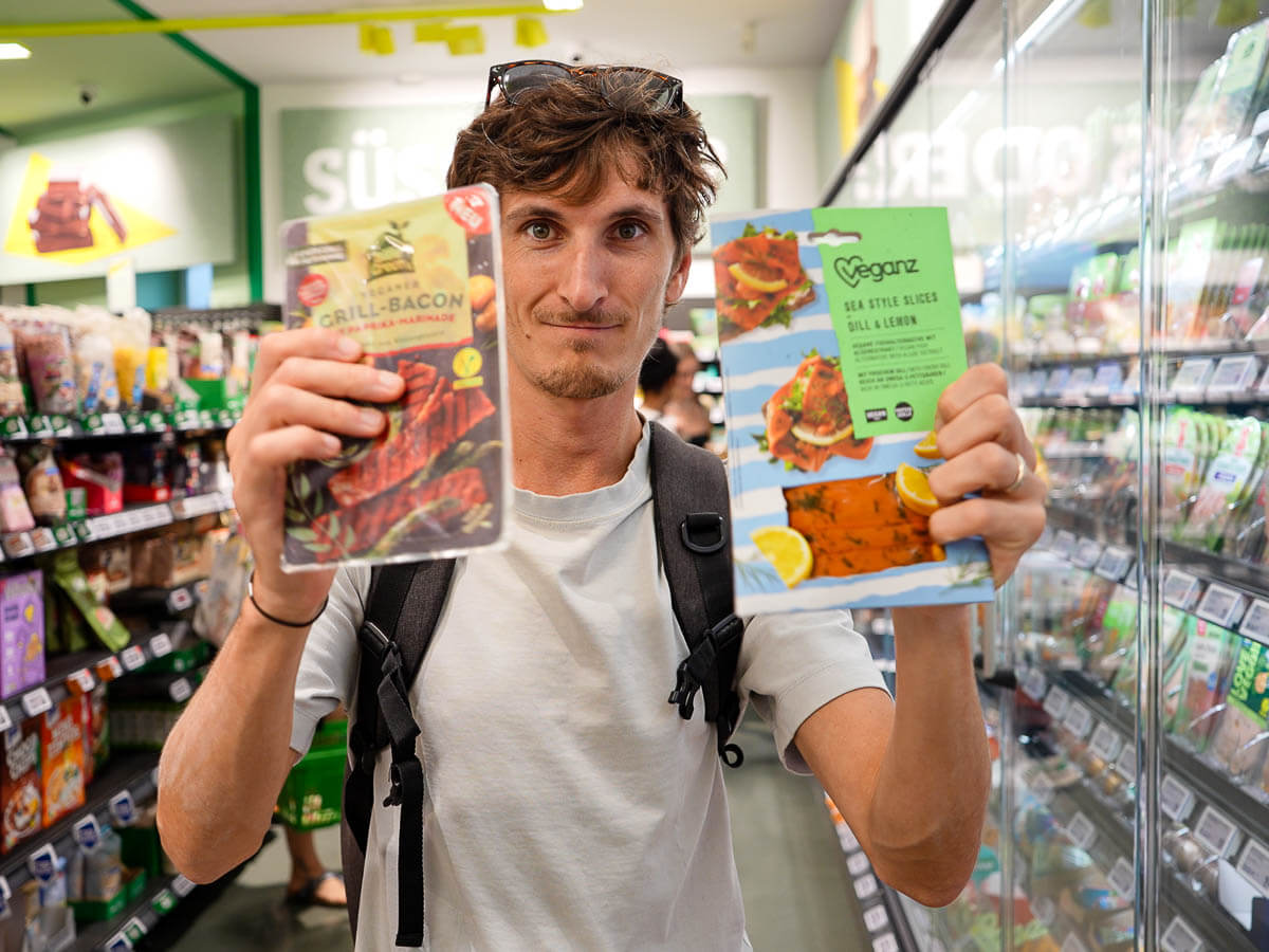 A person standing in a grocery store aisle, holding two different vegan products and looking directly at the camera.