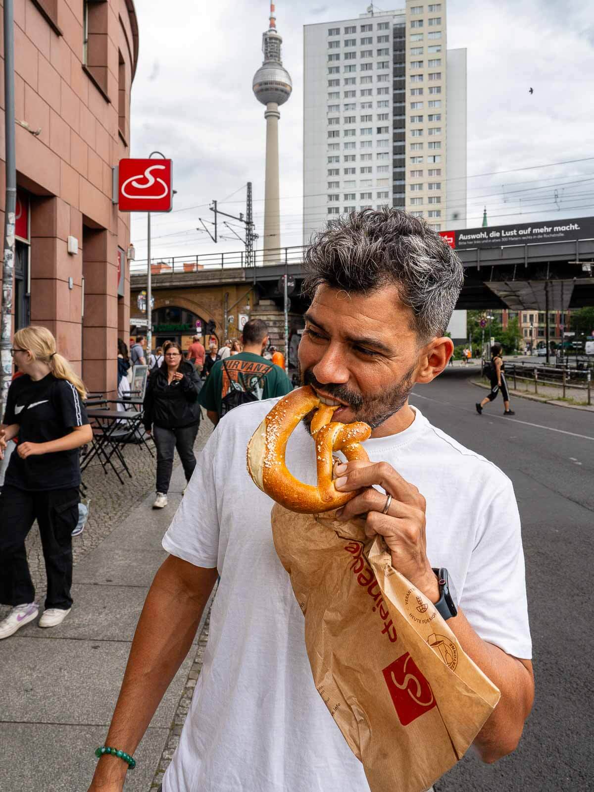 A man enjoying a freshly baked pretzel, taking a bite while standing near a busy street in Berlin, capturing a casual moment in the city.