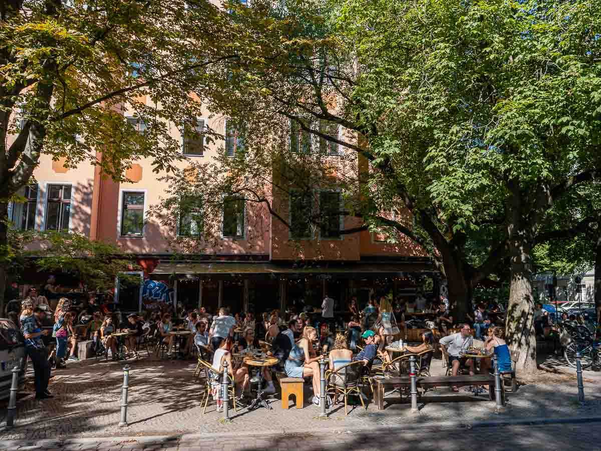 A bustling outdoor scene at a popular Berlin café, where people are enjoying their meals and drinks under the shade of trees, with a vibrant atmosphere.