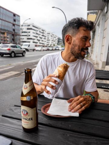 A man sitting at an outdoor table, enjoying a vegan doner with a bottle of vegan beer beside him.