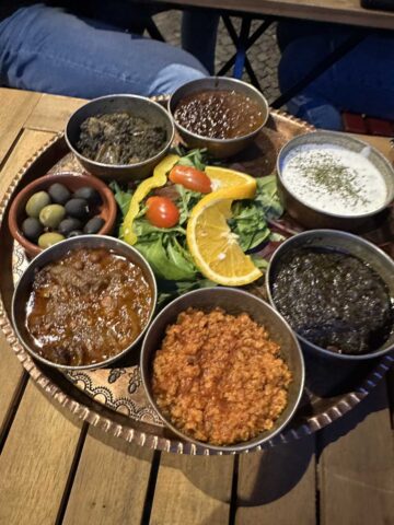 A traditional Persian platter with various vegan dips, olives, greens, and vibrant garnishes, all arranged in small bowls on a large metal tray.