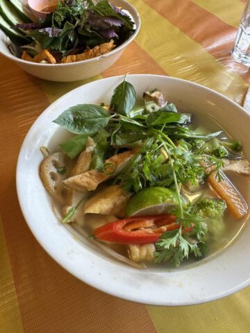 A vibrant bowl of vegan pho filled with fresh herbs, tofu, lime, and sliced chili peppers, served in a white bowl on a wooden table.