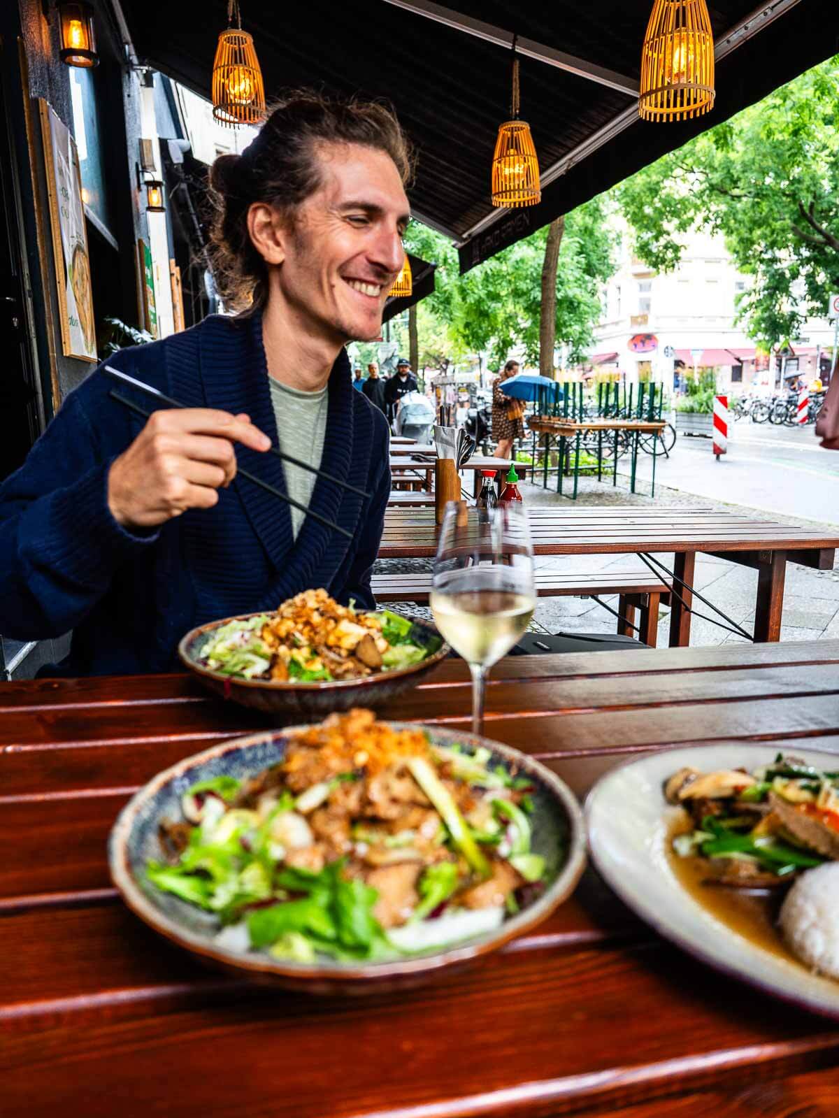 A person enjoying a meal at an outdoor table, smiling while eating a colorful vegan dish with salad, seated at a cozy restaurant in Berlin.