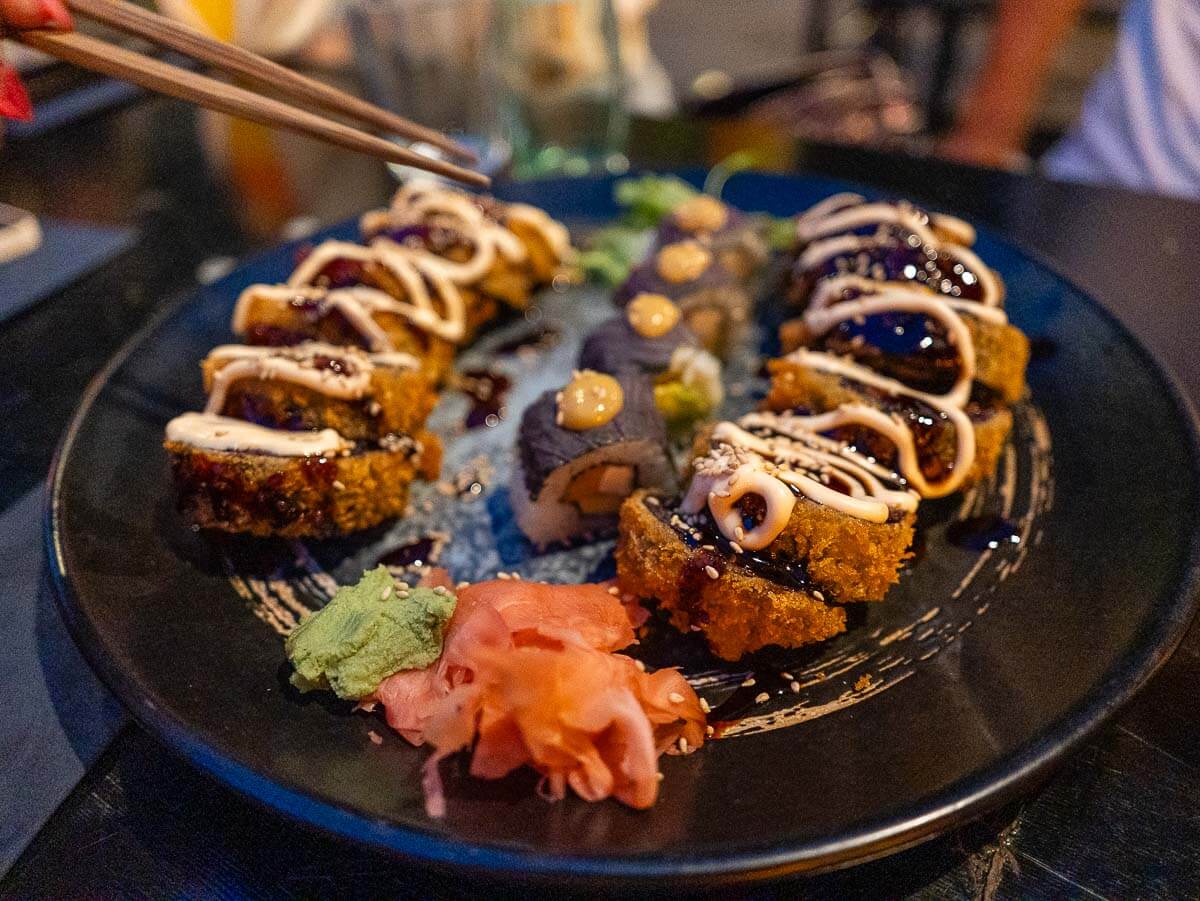 Another angle of the vegan sushi roll, showcasing its vibrant toppings and detailed presentation, surrounded by fresh garnishes on a black plate.