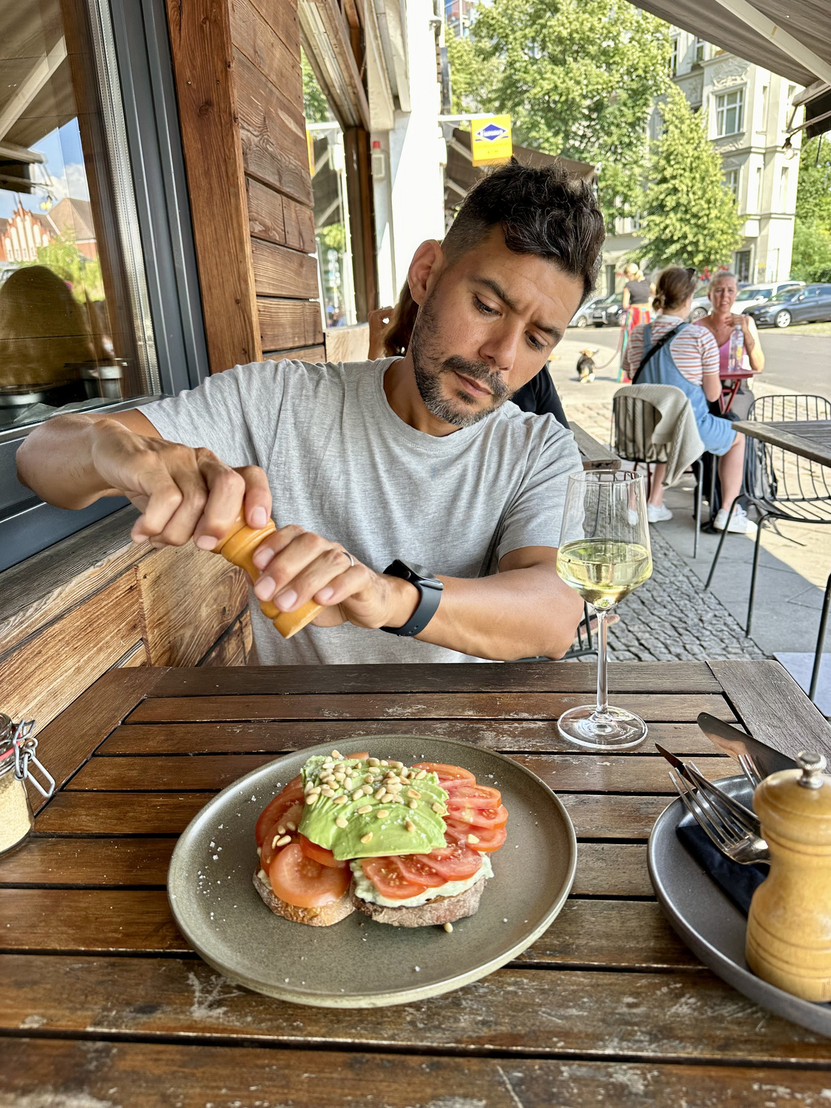 A man sitting at an outdoor café table, seasoning his vegan avocado toast with freshly ground pepper, with a glass of white wine and a relaxed atmosphere.