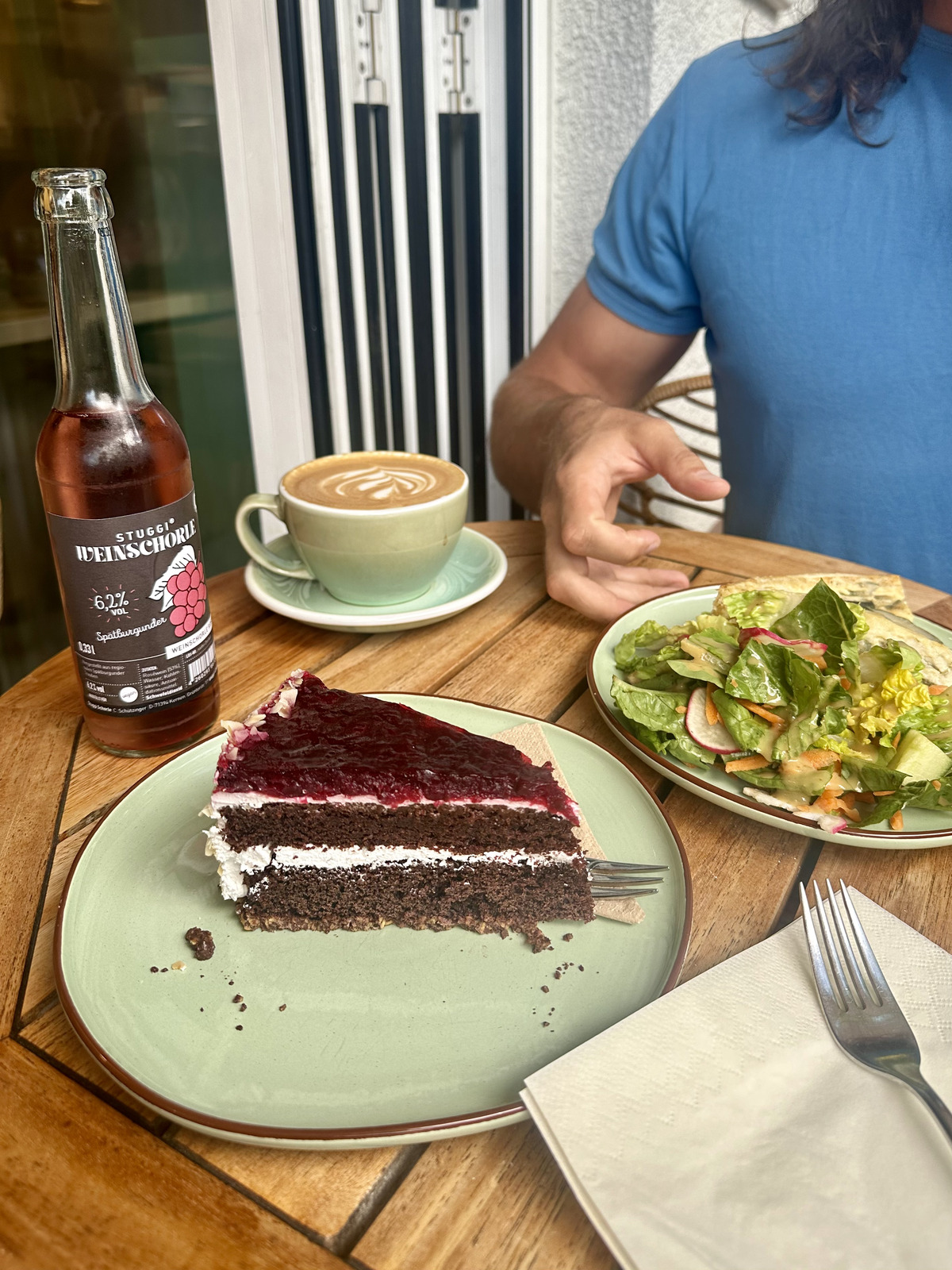 A slice of rich vegan chocolate cake served on a green plate alongside a bottle of kombucha and a cup of coffee, with a fresh salad visible in the background.