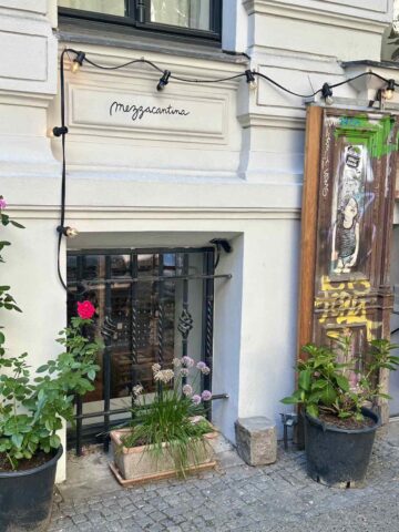 The charming exterior of Mezzacantina, a quaint Berlin restaurant with a wooden sign, potted plants, and a welcoming entrance.