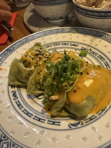 A close-up of a plate of vegan dumplings topped with a creamy sauce and garnished with green onions and sesame seeds, served on a patterned dish.