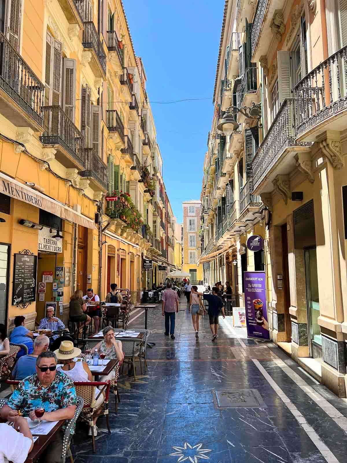 A bustling pedestrian street in Málaga’s historic center, filled with people walking on shiny marble pavement, with colorful decorations overhead.