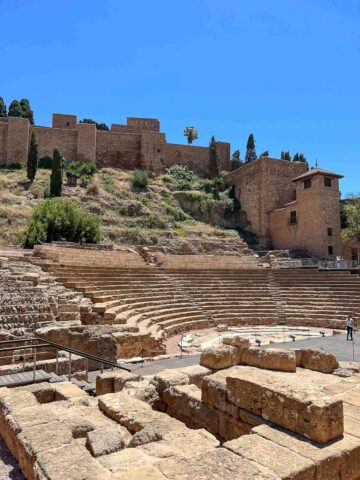 The Roman Theater in Málaga with a crowd of people admiring the ancient ruins, with the Alcazaba fortress visible in the background.