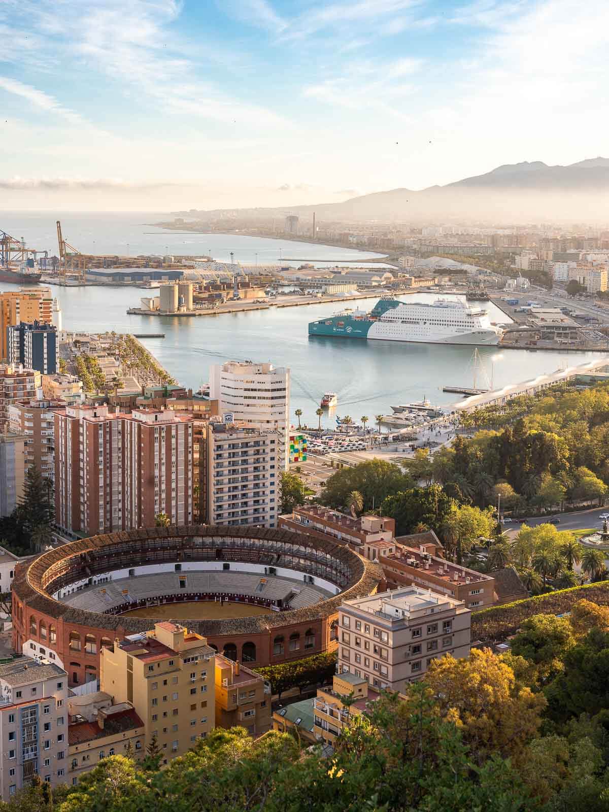 A panoramic view of Málaga’s port and La Malagueta beach, with the Plaza de Toros bullring in the foreground and the mountains in the background under a bright blue sky.