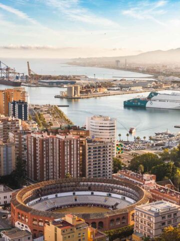 A panoramic view of Málaga’s port and La Malagueta beach, with the Plaza de Toros bullring in the foreground and the mountains in the background under a bright blue sky.