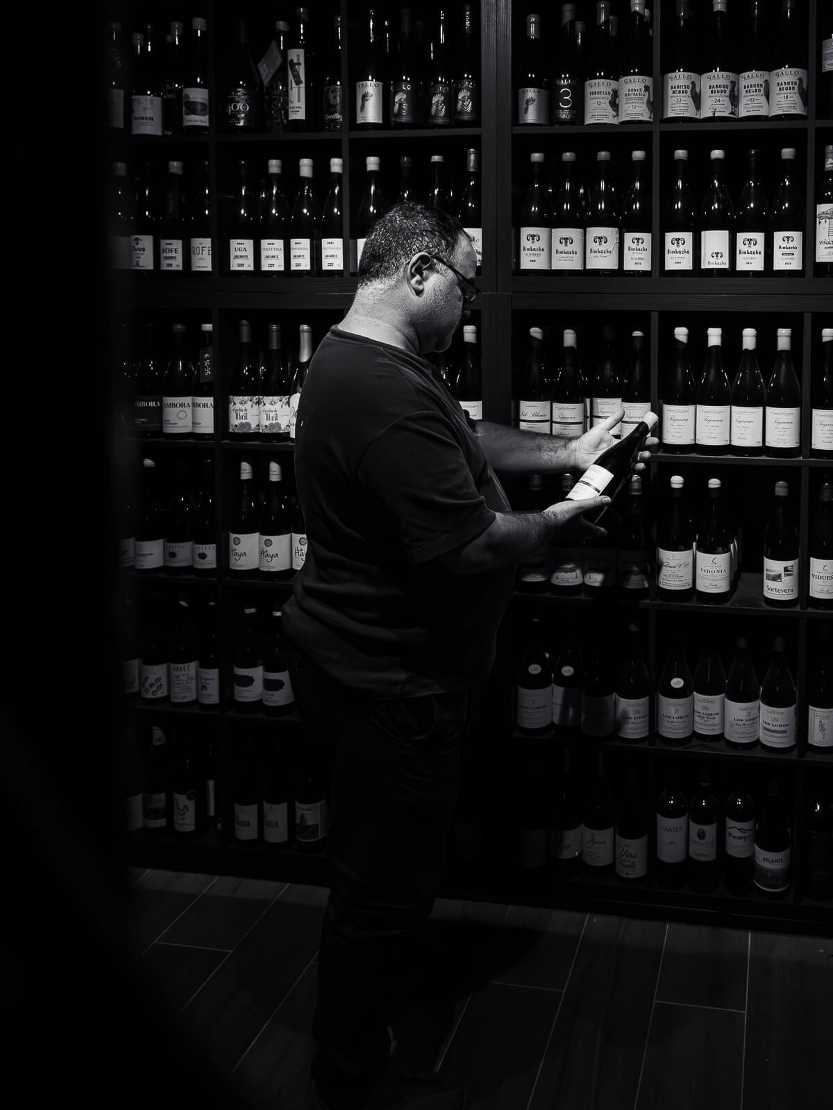 Jacinto selecting a bottle of wine from the shelves in Vinoteca Con Pasión, surrounded by an extensive collection of Canarian wines in a dimly lit ambiance.