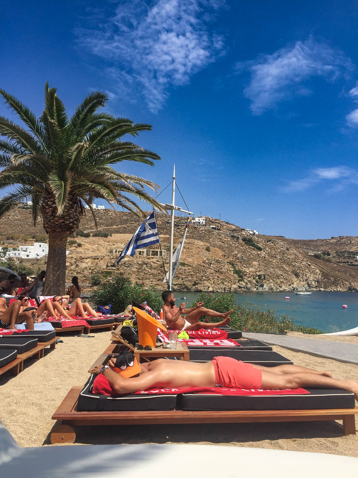 Relaxing at a beach club in Mykonos, with palm trees, the Greek flag, and turquoise sea in the background.