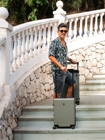 Stylish traveler posing on stone staircase with LEVEL8 Voyageur suitcase set in olive green.

