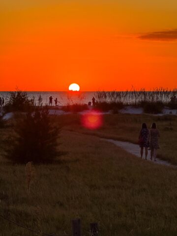 People walking along a sandy path toward the beach during a vibrant orange sunset on Anna Maria Island, Florida.